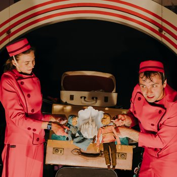 A man and a woman in red jumpsuits and hats are holding up two dolls in front of an open vintage suitcase. There is an arched backdrop behind them.