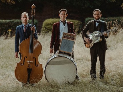 Photograph of the three members of the Leeds City Stompers with their instruments; a double bass, drum kit, guitar and washboard.