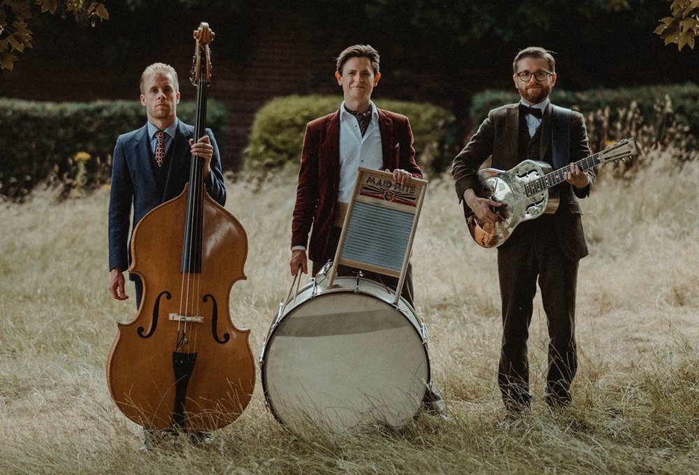 Photograph of the three members of the Leeds City Stompers with their instruments; a double bass, drum kit, guitar and washboard.