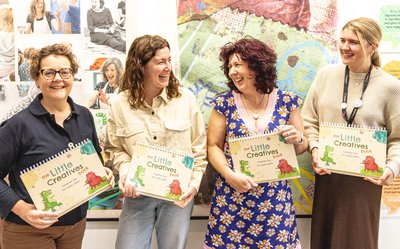 Four women laughing stood in front of an exhibition holding the Little Creatives book.