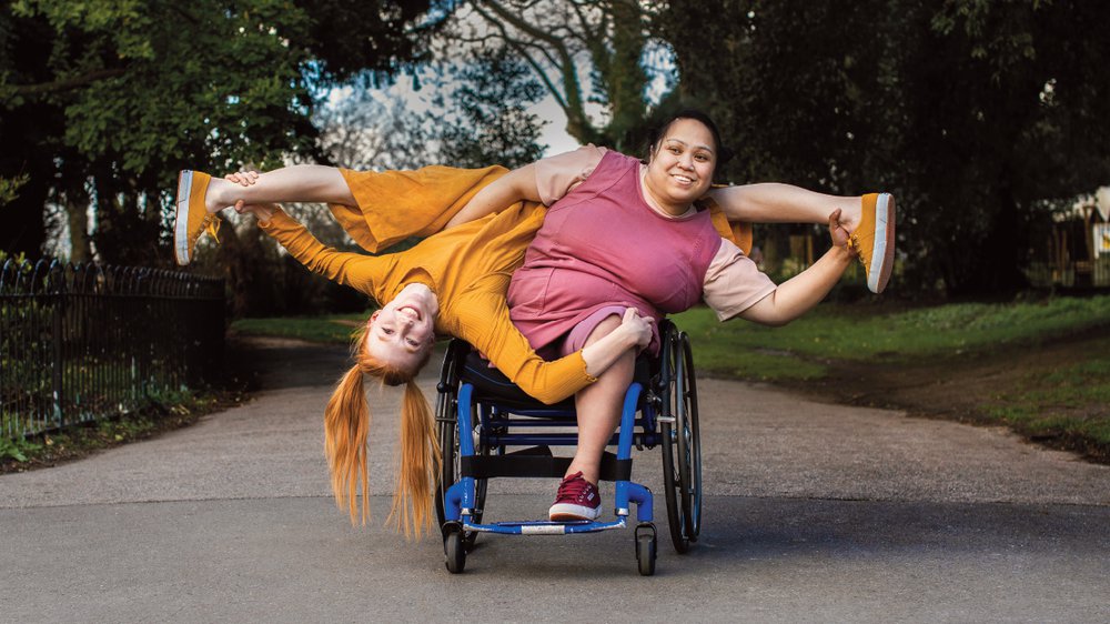 a curvy brown skinned filipino female wheelchair dancer, with an above right knee amputation sat in a manual wheelchair, she leans to the left, her left hands holds the ankle of another woman whose leg rest on her shoulders. She wears a plum coloured dress. Her right arm is clasped around the waist of a white woman with ginger hair who hangs upside down with her legs in a split. The woman with ginger hair holds her left ankle and has her left hand on the first woman’s left knee, she wears an ochre jumpsuit. They look out at viewer smiling. Behind them is a grey asphalt path going off into the distance, they’re framed by trees and shrubbery a blue sky peaking out from between the leaves.