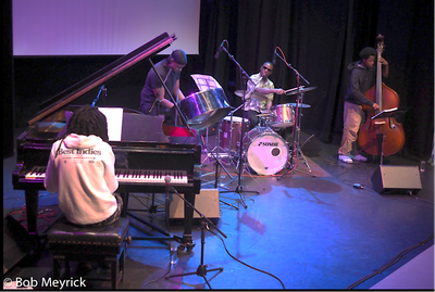 Photograph of seated piano player and jazz quartet on stage