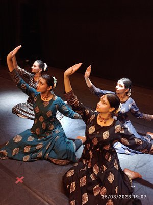 Four female dancers sit on a stage with their arms raised in a classical Indian Kathak style, wearing classical Indian dress.