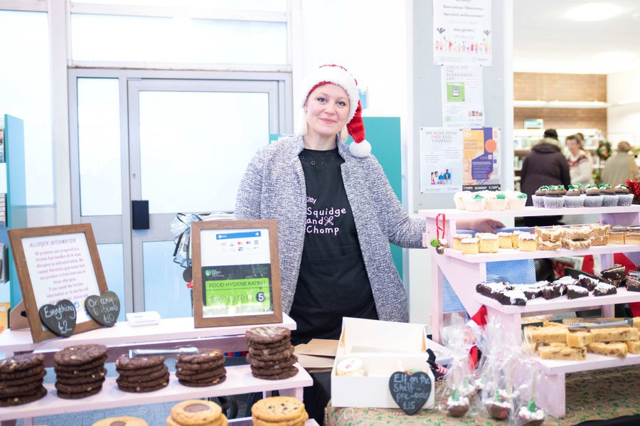 Smiling lady in Santa hat behind stalls at library Christmas market