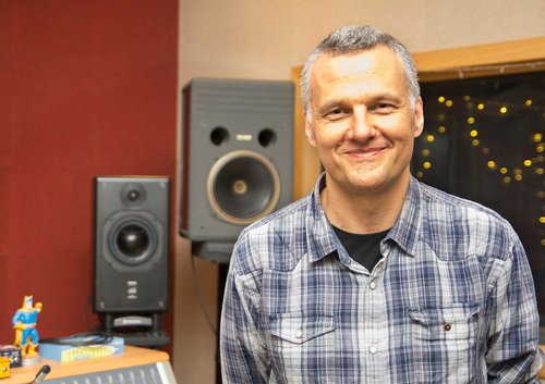 A man in a checked shirt smiling in a recording studio.