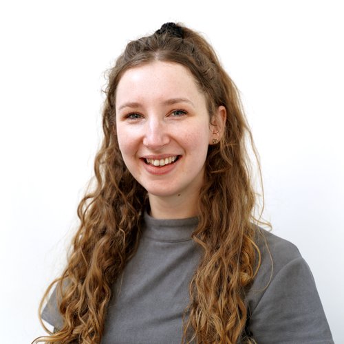 Female with long brown curly hair, smiling to the camera. Wearing a grey top, standing against a white background.
