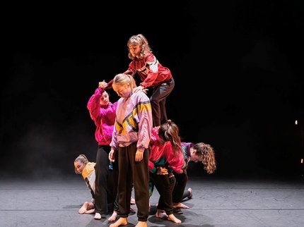 Six young dancers in bright jackets lifting one dancer