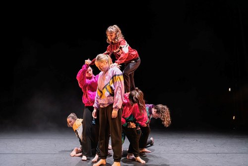 Six young dancers in bright jackets lifting one dancer
