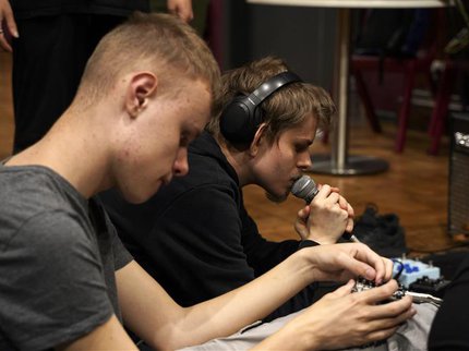Two boys sat on the floor. One is wearing headphones and speaking in to a microphone.