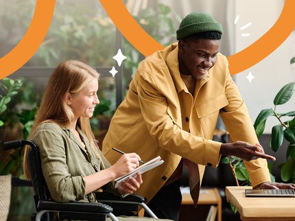 A smart young black man in casual clothing and a beanie hat smiles and points at a laptop advising a white female in a wheelchair with long light brown hair.
