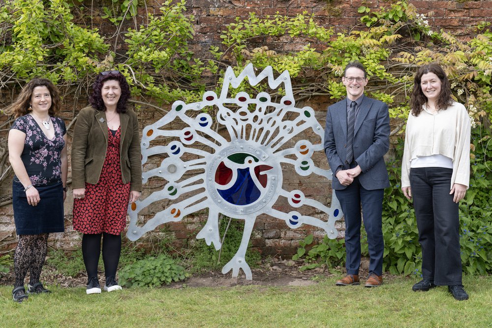 Four people stand next to the sculpture of the metal peacock with colourful glass feathers