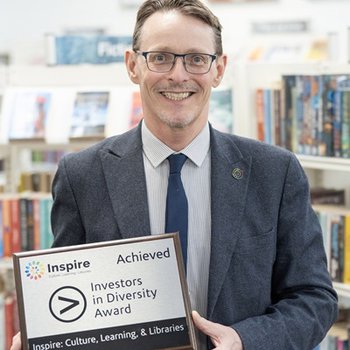 Peter Gaw has short brown hair, wears glasses, a light blue shirt striped shirt with a dark blue tie and jacket. Peter smiles whilst holding a silver plaque on a dark brown wooden mount with the words Investors in Diversity Award.
