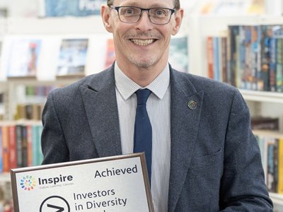 Peter Gaw has short brown hair, wears glasses, a light blue shirt striped shirt with a dark blue tie and jacket. Peter smiles whilst holding a silver plaque on a dark brown wooden mount with the words Investors in Diversity Award.