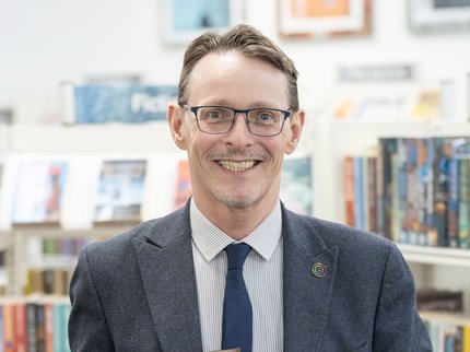 Peter Gaw has short brown hair, wears glasses, a light blue shirt striped shirt with a dark blue tie and jacket. Peter smiles whilst holding a silver plaque on a dark brown wooden mount with the words Investors in Diversity Award.