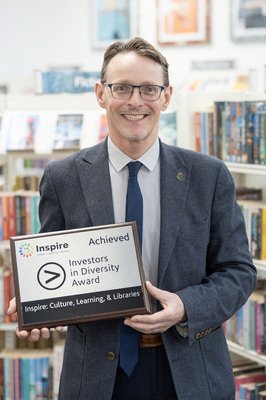 Peter Gaw has short brown hair, wears glasses, a light blue shirt striped shirt with a dark blue tie and jacket. Peter smiles whilst holding a silver plaque on a dark brown wooden mount with the words Investors in Diversity Award.