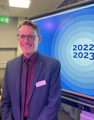 Peter Gaw, Inspire Chief Executive, a man with brown hair stood in a suit with a plum coloured shirt in front of a screen with blue circles and 2022 2023 in the middle