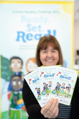 A smiling, female librarian holding up three of the pull-out folders that children will get when they join the challenge