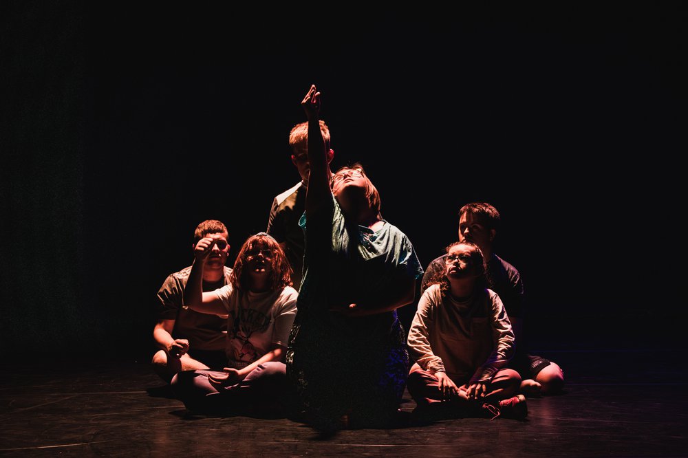 A dark image of 6 young people sat on a stage. The front person is raising their arm.