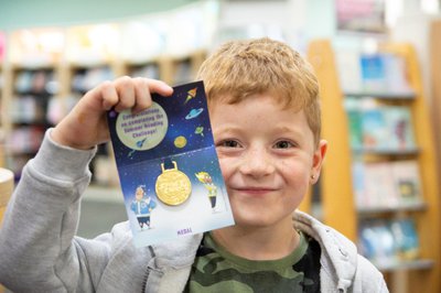 A young boy, smiling, holds up a Summer Reading Challenge medal.