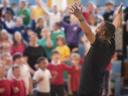 Children taking part in singing event by a man singing with uplifted arms