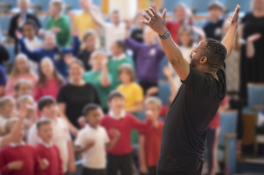 Children taking part in singing event conducted by a man singing with uplifted arms