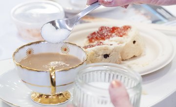 Photograph of a cup of tea with someone adding a spoon of sugar with a scone on a plate next to the cup and saucer.