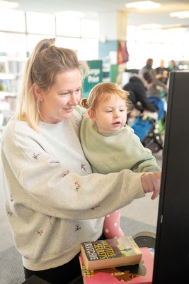 Blond lady holding child in the library and using the self service screen to borrow books