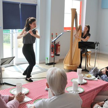 Two musicians are playing to a group of people sat around tables. One is playing a flute, the other is playing a harp.