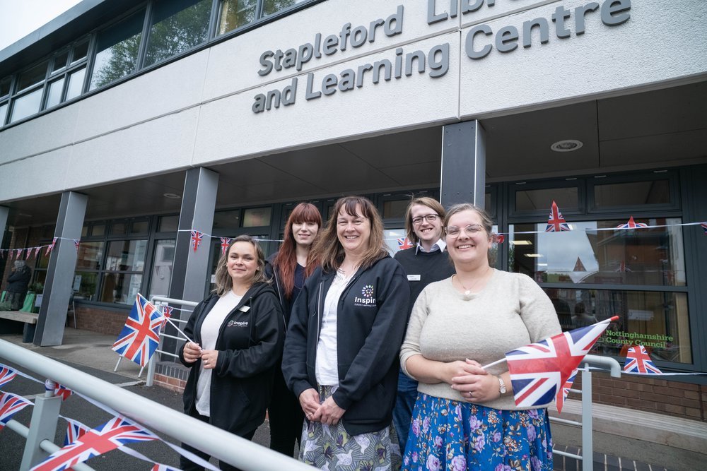 Five members of staff stood in front of Stapleford Library, smiling and holding union jack flags