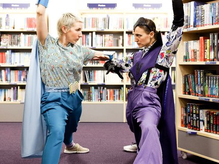 Two actors in superhero outfits, posing in a library.
