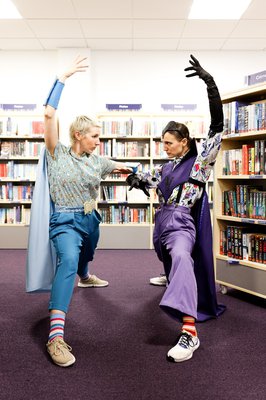 Two actors in superhero outfits, posing in a library.