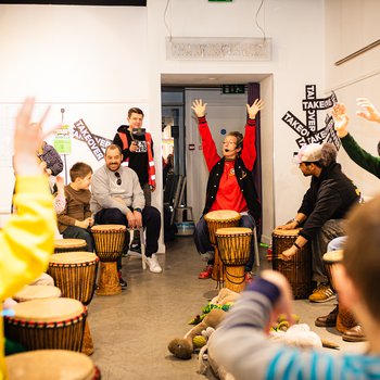 A group of people sit in a circle with a woman in a red jumper playing drums