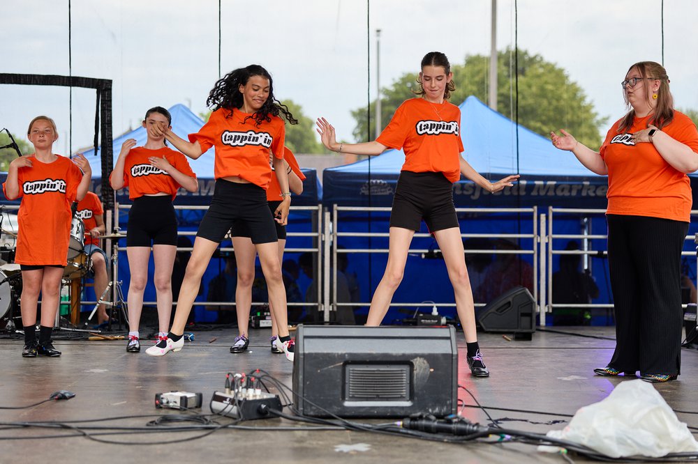 Five young dancers on stage wearing orange T shirts jumping in the air.