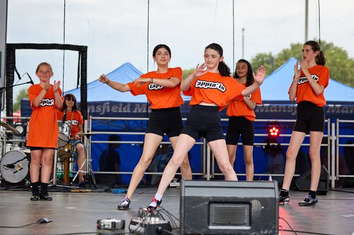 four dancers on stage in orange t-shirts, black shorts and black tap shoes with a drummer behind them.