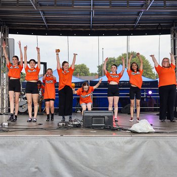 9 young dancers on stage wearing orange Tshirts raising both arms above their heads.