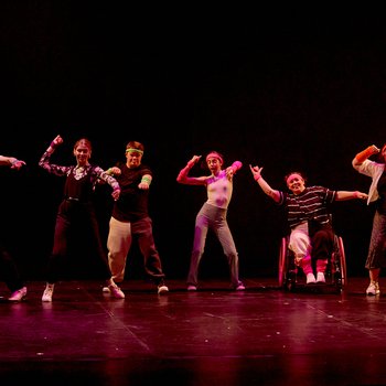 Six young dancers performing on stage smiling with their right arm raised. One dancer is in a wheelchair.