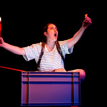 A dark haired girl, in a striped t-shirt, with straps of backpack over her shoulders stares open mouthed at crisp she is holding high in her left hand.