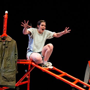 A dark haired girl in striped t-shirt and denim shorts crouches on a bright orange climbing frame, arms outstretched.  A dark green wax jacket hangs on the frame next to the girl.