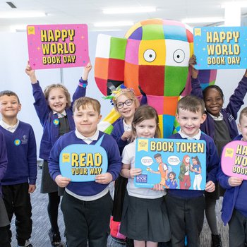 Smiling school children  surround Elmer the elephant, holding signs that say "Happy World Book Day" and "Read Your Way"