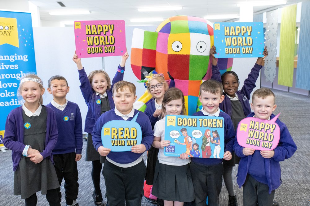 Smiling school children  surround Elmer the elephant, holding signs that say "Happy World Book Day" and "Read Your Way"