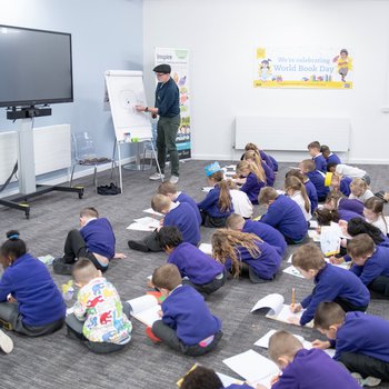 School children sit on the floor, leaning forward drawing watching author Rob Biddulph