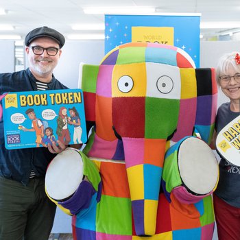 Smiling school children  surround Elmer the elephant, holding signs that say "Happy World Book Day" and "Read Your Way"