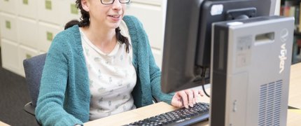 woman with brown hair and glasses sat clicking a mouse and typing on a computer