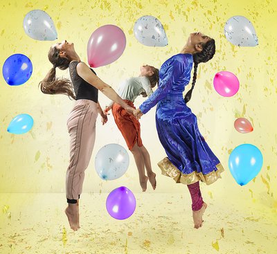 Three dancers jump in the air whilst holding hands in a circle surrounded by balloons.