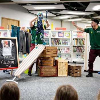 An actor pupeteer holds Blue pupper, Bo, above a ironing board leaning against boxes like a slide.  A second actor holds his hand aloft in triumph.