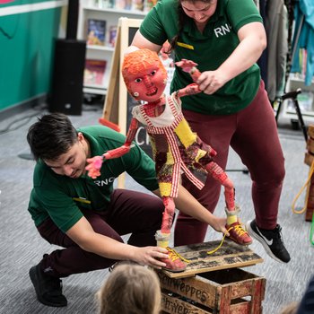 A bright red rag doll style puppet, Little Red, is balanced by two puppeteers on a rickety wooden box.