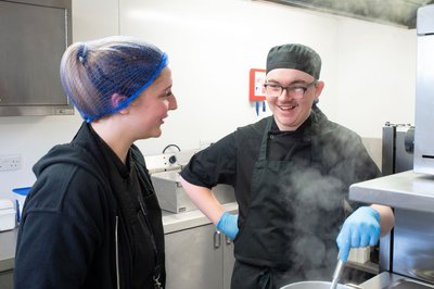 A catering student and teacher chatting whilst wearing chef clothing in a kitchen.