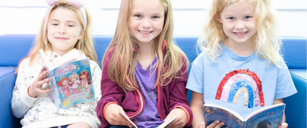 three young girls sat smiling on a sofa with an open book in their hands