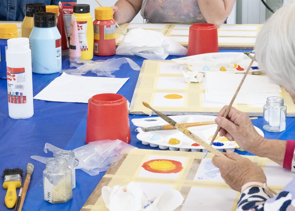 An older person's hands using paint brushes, surrounded by tubes of paint and an artist's palette.
