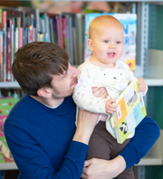 a man in a blue jumper is smiling and looking at the toddler he's holding. toddler has ginger hair and is holding a picture book